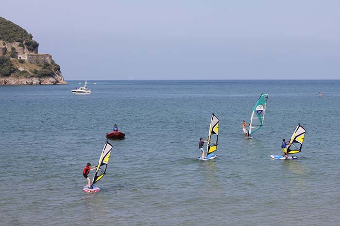 Varios niños practican con sus tablas y velas de windsurf mientras un monitor les vigila desde una lancha